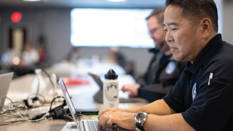 Photo of CISA employees working by laptop and tv monitors