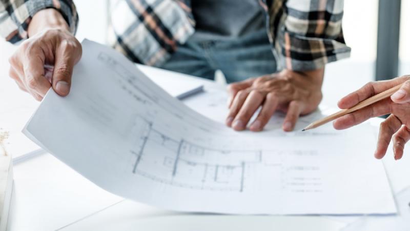 Two sets of hands holding and pointing to blueprint plans on a desk