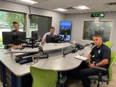 Two radio DJs sit at a recording studio across the table from the CISA Region 8 Regional Director. All are holding headphones that they will put on when they go on air.