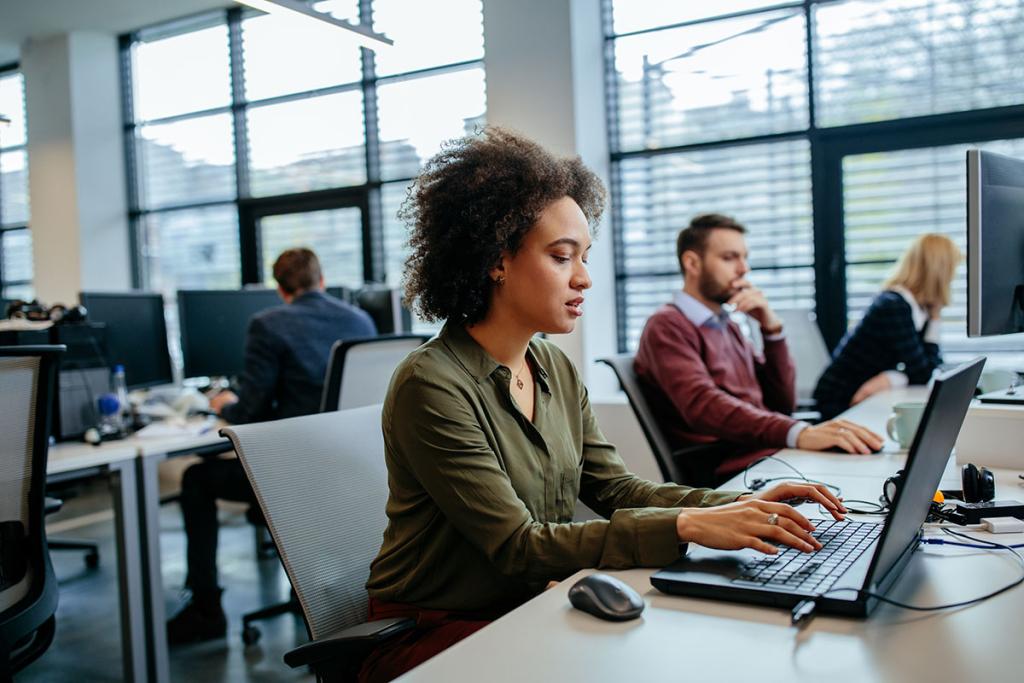 Diverse woman typing on laptop with 4 other colleagues in background in an office.