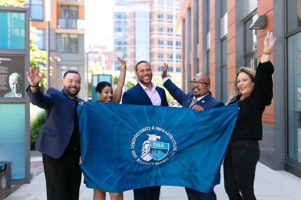 Photograph of diverse CISA staff holding CISA flag and waving in front of CISA HQ.