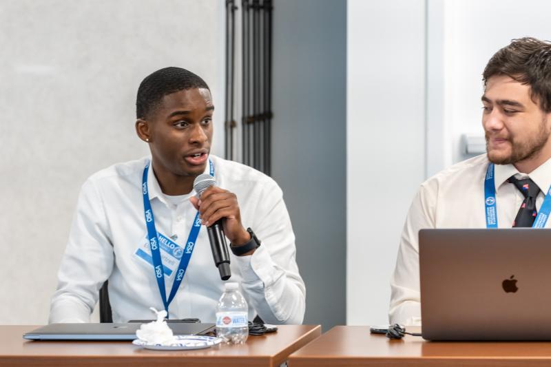 Two male CISA interns talking, one in front of a computer, at a meeting.